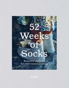 Light gray edged book with "52 Weeks of Socks" title in middle; "Beautiful patterns for year-round knitting" written underneath, "Laine" written on bottom; Close up on feet of child wearing blue knitted socks standing on dark gray rock surface; Bottom of scalloped skirt above socks in blue, yellow, and pink pattern