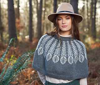 Image of woman standing in the woods wearing tan hat and knitted gray and white shawl around shoulders. Shawl is deep gray background with white feathers coming down from collar