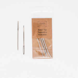 Brown and clear package of bent tip tapestry needles laid out on white background with two needles beside