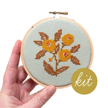 Load image into Gallery viewer, A hand holds a mini embroidery hoop containing a cross-stitch kit design of yellow-orange flowers with brown leaves on a pale blue-gray fabric. A gold-toned metal clamp is visible at the top, and a gold circular sticker with the word 'kit' is in the lower right corner.