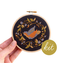 Load image into Gallery viewer, A completed cross-stitch embroidery piece in a wooden hoop, held by a hand against a white background. The design features a stylized orange and gray bird inside a circular wreath of gold and gray leaves with small orange berries on a black fabric background.