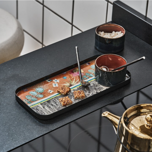 Image of rectangular food tray on gray table top. Tray features colorful images of plants and flowers in blues, oranges, pinks, yellows, and gray. Gray and copper colored mug sticks with spoon sticking out on tray. Second mug sits to right of tray on table with gold colored tea pot in front of tray on table top