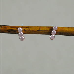Light purple stud earrings with three small circles in a line going down. Studs are piercing long piece of wood on green background