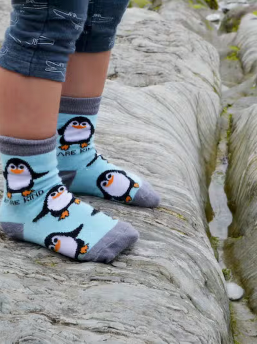 Kid wearing Sky blue socks with grey cuffs, heels and toes. Black and white penguins with orange beaks line the socks. The name Bare Kind is written in black under the first row of penguins.