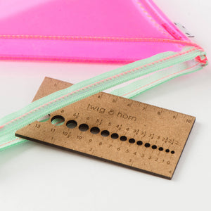 Brown needle gauge that reads "twig & horn" half out of clear and pink zipper pouch with light green zipper