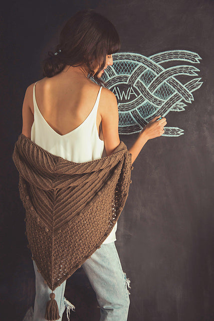 Image of back of woman wearing white tank top and brown shawl drawing on chalkboard the word 
