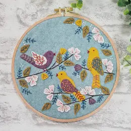 Tan colored embroidery hoop with light blue background fabric. Three birds in yellow, purple, and blue sit in middle on small branches. Pink and white flowers, green and blue leaves, and blue decorative sprigs come off of each branch and surround the birds
