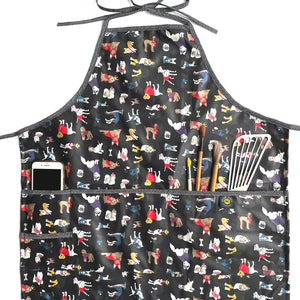 Black apron on white background. Images of dogs wearing clothes in red, purple, blue, and yellow cover the apron. Edges of apron in white and black pattern. Phone, paint brushes, and metal spatula stick out of pocket running along middle of apron