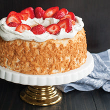 Load image into Gallery viewer, Image of cake on white platter with gold base on dark wood table with black wall background. Cake is light brown and unfrosted around edges. Top of cake is covered in a ring of white whipped cream and bright red strawberry halves. Laying beside platter on table is light blue and white kitchen towel