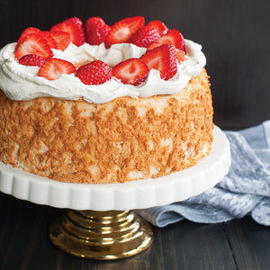 Image of cake on white platter with gold base on dark wood table with black wall background. Cake is light brown and unfrosted around edges. Top of cake is covered in a ring of white whipped cream and bright red strawberry halves. Laying beside platter on table is light blue and white kitchen towel