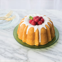 Load image into Gallery viewer, Light brown baked bundt cake with white icing dripping down sides and raspberries piled in middle; Sprig of green garnish sticking out of raspberries; Cake sitting on light green striped glass plate on top of gray and white countertop; 2 clear plates with two gold colored forks to the left of cake