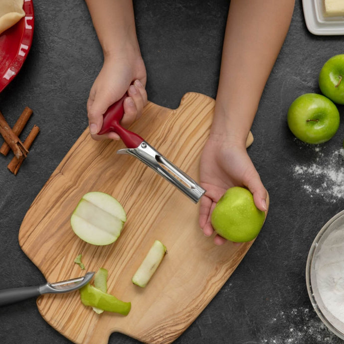 Image of person about to de-core a green apple over light wood cutting board. Green apples, flour, cinnamon sticks, and a peeler are all spread out around the cutting board and everything rests on a dark gray countertop