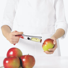 Load image into Gallery viewer, Person in white apron against white background holding red apple in one hand and red and stainless steel apple corer in other. Apple core sits in middle of apple corer and three red apples sit on white countertop in front of person