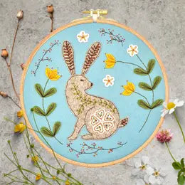 Light tan embroidery hoop on gray background with sprigs of wild flowers around. Rabbit in brown and white sits in middle of hoop with small ferns and a four leaf clover covering its back. Sprigs of wildflowers in yellow, green, pink, and white, surround the rabbit in the hoop