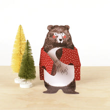 Load image into Gallery viewer, Open example of bear hugs notecard sits in front of two small plastic Christmas trees on wooden table. Bear is wearing red cardigan with white polka dots with its arms out as it blushes