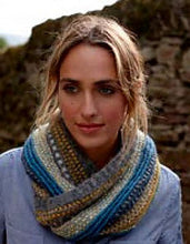 Load image into Gallery viewer, Image of woman wearing blue shirt and knitted cowl in front of stone wall. Cowl is in shades of yellows, blues, and grays and is twisted around the woman&#39;s neck as she looks off to side