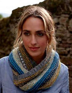 Image of woman wearing blue shirt and knitted cowl in front of stone wall. Cowl is in shades of yellows, blues, and grays and is twisted around the woman's neck as she looks off to side