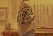 Load image into Gallery viewer, Image of woman wearing knitted sweater standing to the side in front of long wooden table and wall covered in macrame. Sweater in tan and black with tribal pattern running across