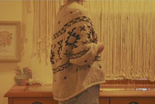 Image of woman wearing knitted sweater standing to the side in front of long wooden table and wall covered in macrame. Sweater in tan and black with tribal pattern running across
