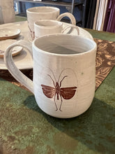 Load image into Gallery viewer, Off white clay mug with image of flying bug on front sitting on green metal table