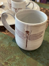Load image into Gallery viewer, Off white mug with image of brown dragonfly on front sitting on green metal table