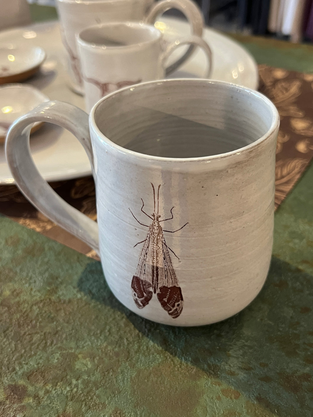 Off white clay mug with image of brown flying bug on front sitting on green metal table