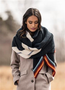 A brunette woman walking with her eyes down in a beige coat wrap in a knitted shawl of navy, white, gray and light blue, red and orange stripes.