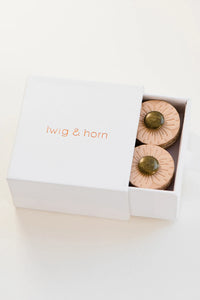 Small white box on white background; Box reads "twig & horn" in bright orange across middle of box; Box is partially open to reveal two round post buttons; Buttons are light tan with bronze colored center and flower-like line indentions spraying out from middle 