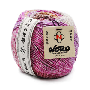 Noro Akari Yarn on white background; Yarn mostly shades of pink, orange, brown, yellow, and tan; Noro tag in tan attached with black string to right side of yarn reads "NORO" "the World of Nature" "MADE IN JAPAN"; Paper wrap around left side of yarn in black and white with Japanese characters running up and down