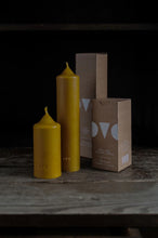 Load image into Gallery viewer, Two mustard colored candles sit unlit on wooden table next to cardboard boxes