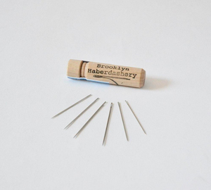 Small light brown wooden needle case on white background with "Brooklyn Haberdashery" written on front on case; 6 small gray stitching needles spread out in fan shape in front of case
