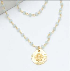 Close up of medallion drop pendant on necklace. Pendant shows image of emblem on it with yellow chain and light blue beads running up from pendant. 