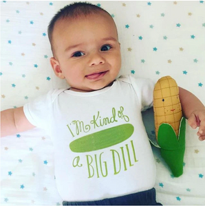 Small toddler lays on starry background with white onesie with pickle on it that reads "I'm kind of a BIG DILL" in green. Child holds stuffed corn toy in crook of arm