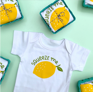 White onesie laid out on green background reads "Squeeze me" in green with image of lemon below. Egg cartons with onesie inside are tied up with string and laid out around onesie