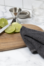Load image into Gallery viewer, Dark grey cloth folded over a wooden cutting board with limes, measuring cup and strainer on counter.