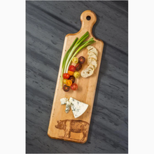 Load image into Gallery viewer, Image of long wooden cutting board on dark gray background. Image of a pig burned into bottom right corner of board with rounded handle at other end. Rest of board covered in cheese, bread slices, tomatoes, and green onions.
