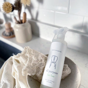 Image of Common Good All Purpose Tea Tree spray bottle resting in small bowl with dish rag on white kitchen counter top with white backsplash behind and wooden bottle brushes in far corner 