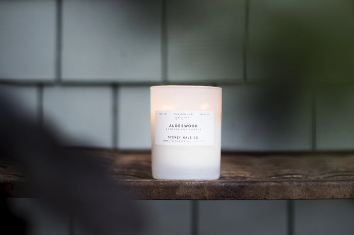 A white candle with a label that reads 'Sydney Hale Co.', placed on a wooden surface with a blurred background.