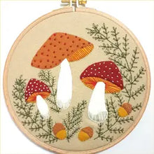 Load image into Gallery viewer, Light tan embroidery hoop with light tan background fabric inside. Three mushrooms in white, orange, and red gather in middle of image, with three yellow and brown acorns below and green ferns all around