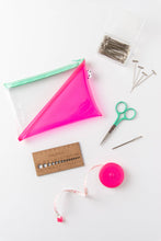 Load image into Gallery viewer, Spread out beginner knitters tool kit on white background. Pink and clear pouch with green zipper, clear plastic container of pins, small green scissors, a tape measurer, and a needle gauge are included in the strewn out items