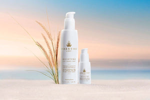 Image of two white bottles with pump tops sitting in the sand in front of the ocean. Bottles reads "Essentiel by Adele, Moisture, Face-Hands-Body"