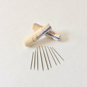 2 light wooden needle casing tubes with stoppers pictured with 9 assorted sized needles laid out on white background; One casing laid out across the other with both reading "9 Assorted Tapestry Needles" in black on blue and white stickers that lay across the stopper tops and connect to casing