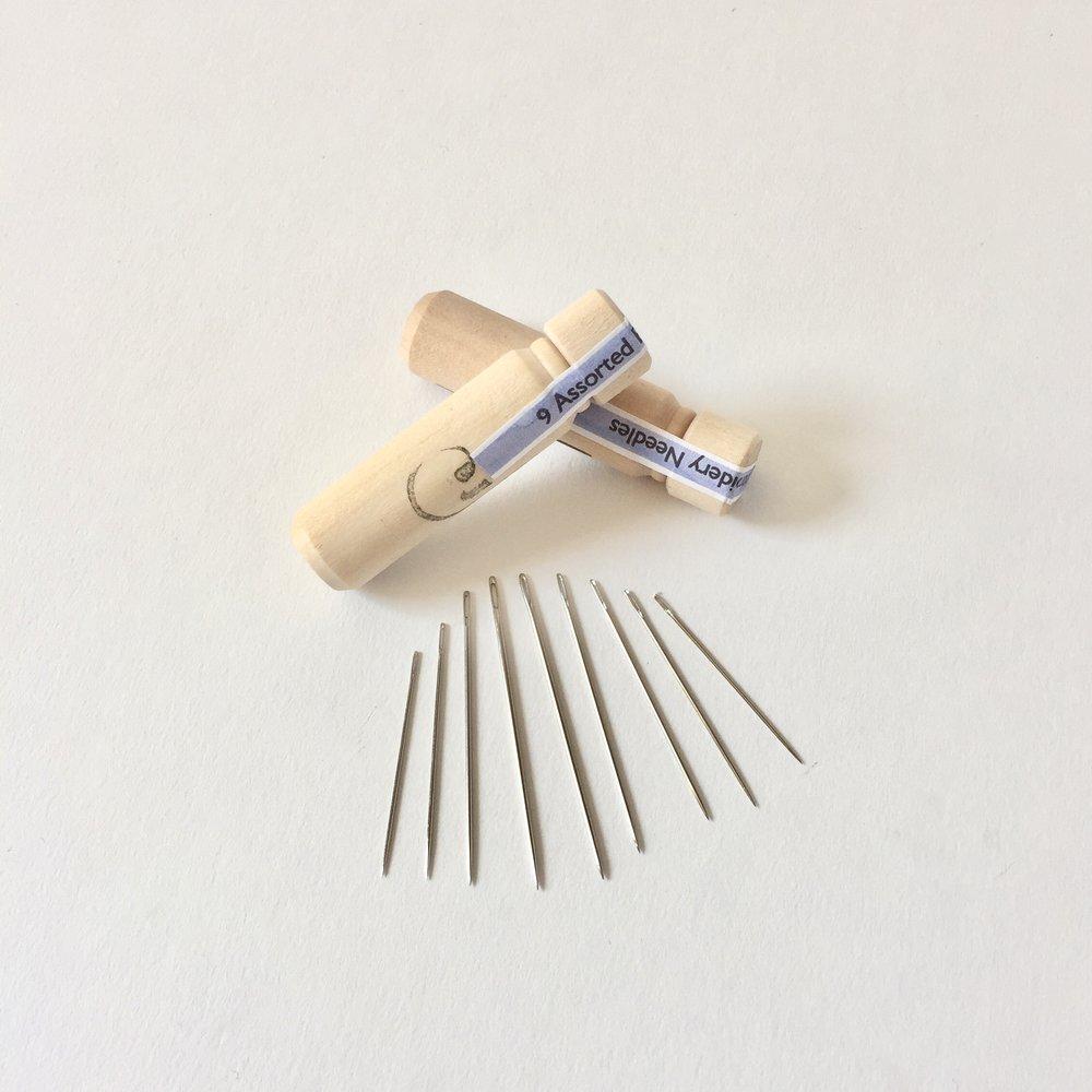 2 light wooden needle casing tubes with stoppers pictured with 9 assorted sized needles laid out on white background; One casing laid out across the other with both reading 