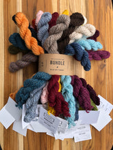 Load image into Gallery viewer, Mini skeins of yarn in 27 different colors cinched together with a Kraft paper strap with white tags hanging from each on a wood tray