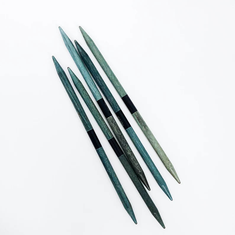 5 Indigo Double Pointed Knitting Needles in smooth driftwood-like shades of blues and greens on white background; small black label wrapped around middle of each 